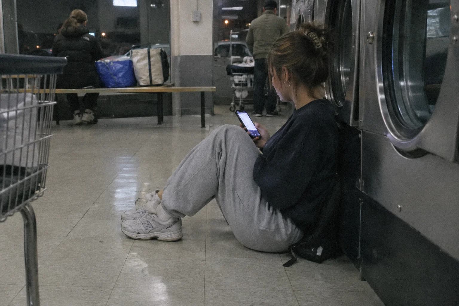 A young woman sitting on the floor of a laundromat at night, her back against a dryer, her lit phone in one hand