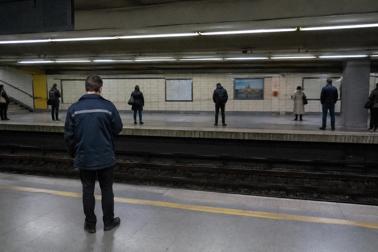 A man standing with his back to the camera on a metro platform, watching the commuters waiting on the opposite platform under strip lights