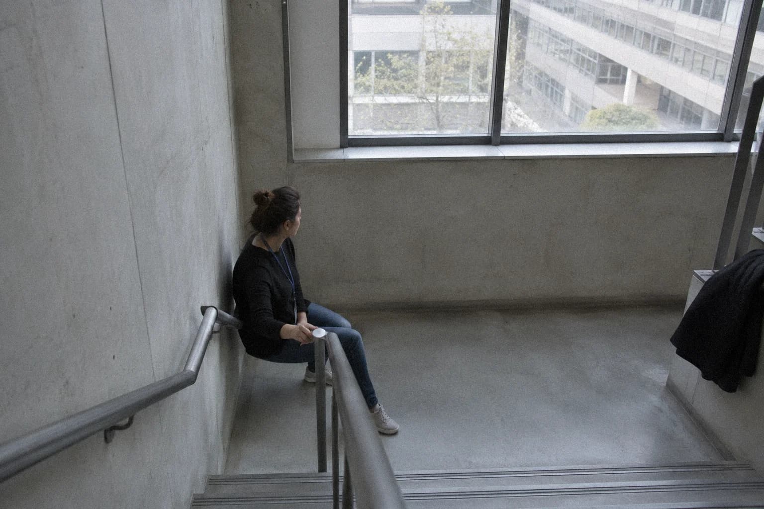 Une femme assise seule sur un palier d’escalier de bureau pendant une pause, un gobelet entre les mains, le regard vers la fenêtre, moment où l’on peut stimuler le nerf vague par la respiration sans que personne le remarque