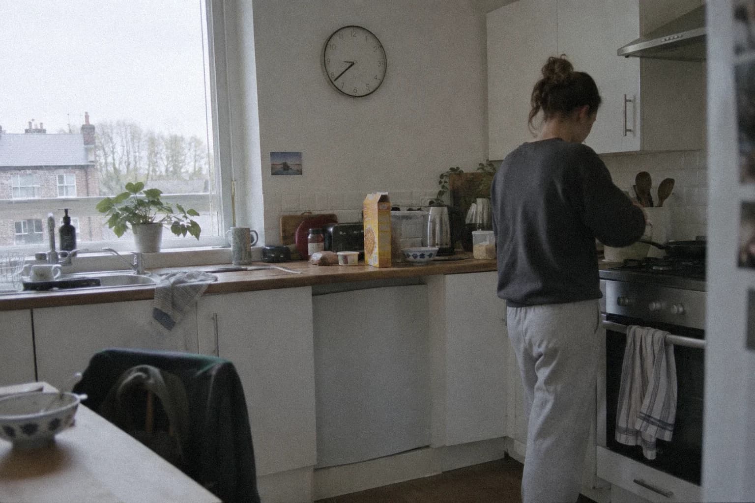 Une horloge murale dans une cuisine d’appartement au petit matin, une personne de dos devant la plaque de cuisson