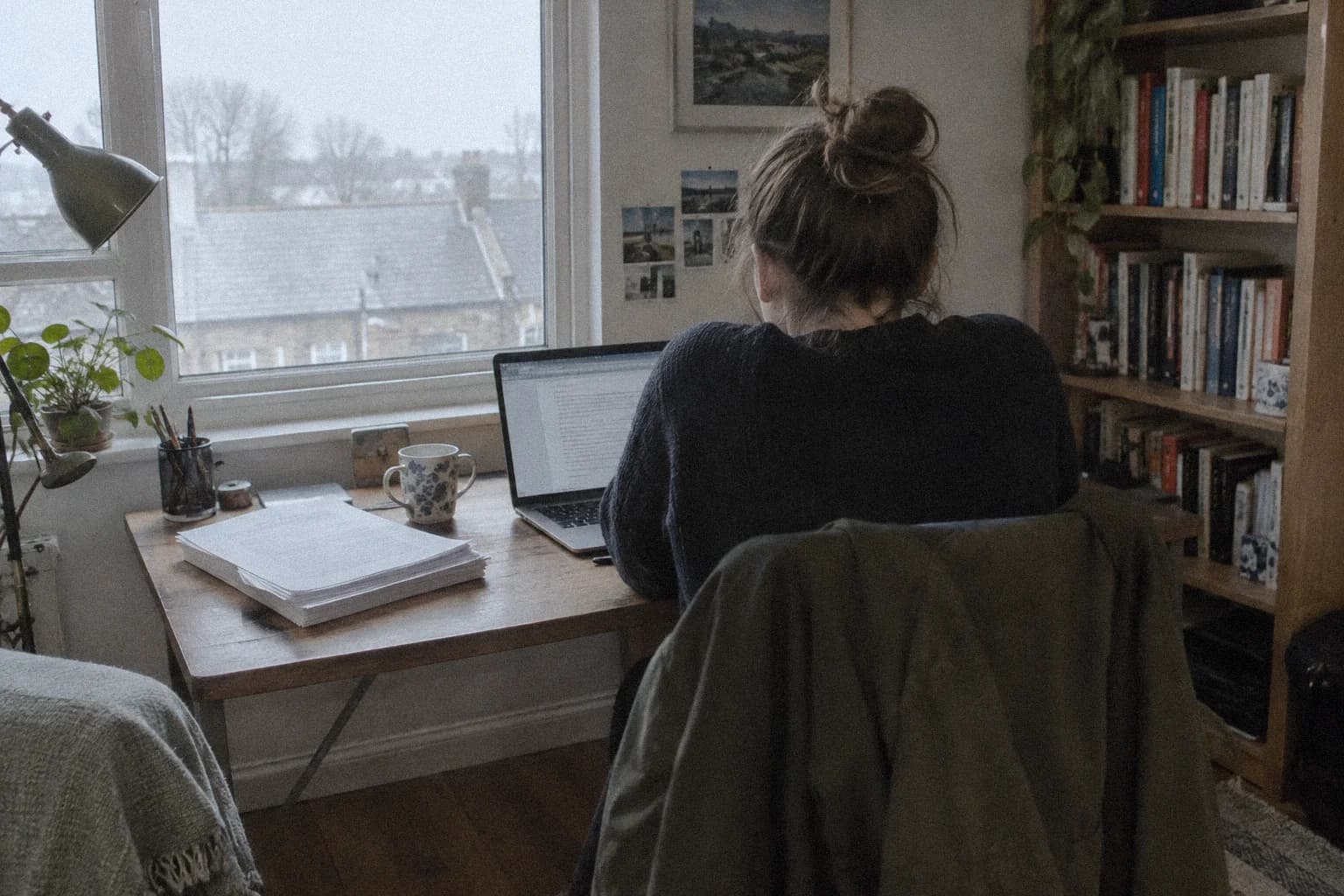 Épaules contractées par le stress : une femme de dos à son bureau un après-midi gris, épaules remontées vers les oreilles devant un ordinateur portable et une pile de papiers