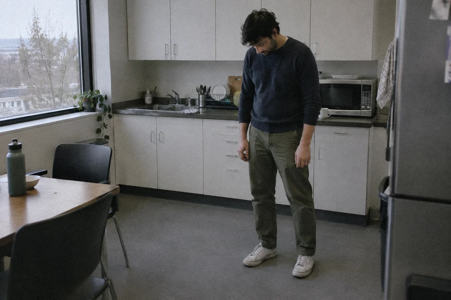 Un homme debout seul dans la cuisine de son lieu de travail pendant une courte pause, les bras pendant le long du corps, le regard vers le sol