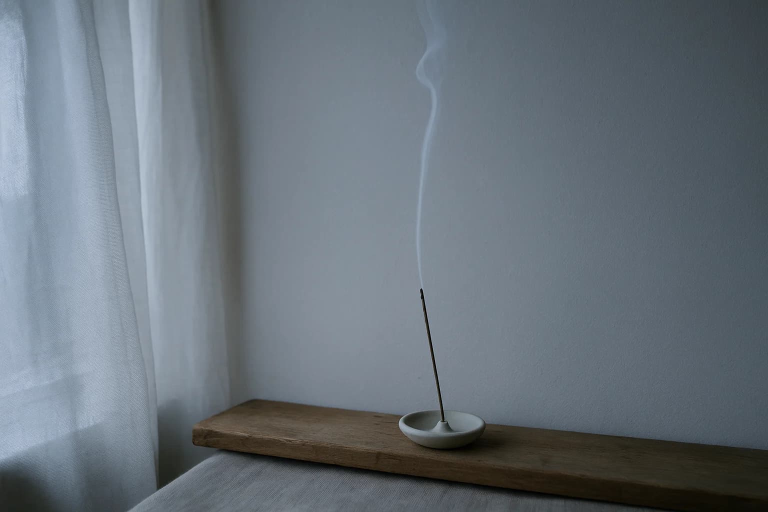 Un mince filet de fumée d’encens qui monte contre un mur clair, sur une étagère en bois