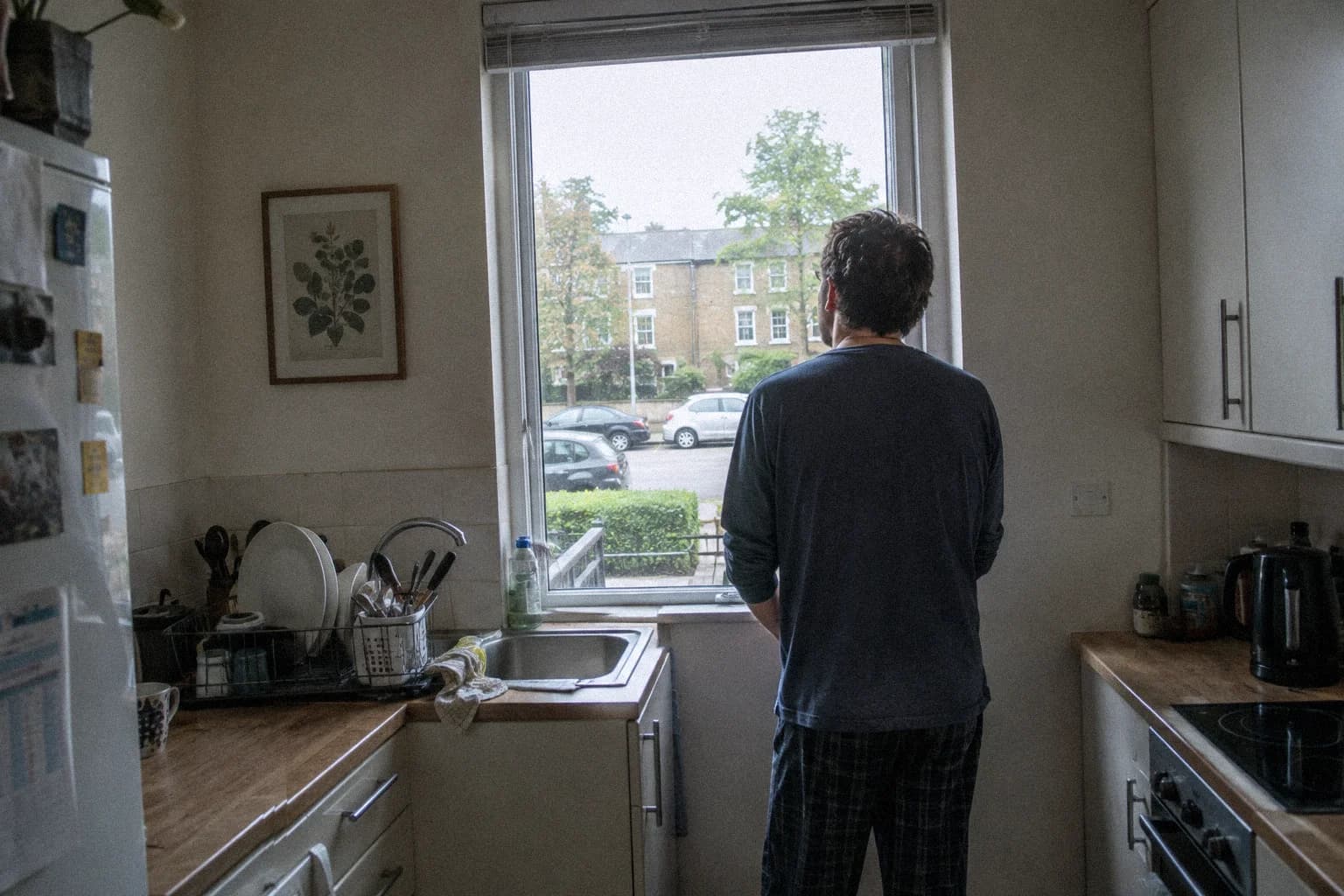 Un homme debout devant la fenêtre de sa cuisine tôt le matin, regardant la rue et les voitures garées, la vaisselle en train de sécher derrière lui