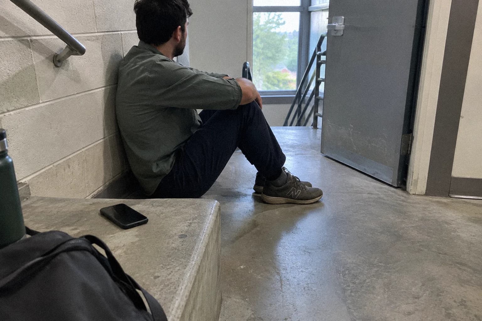 Un salarié assis par terre dans une cage d’escalier pendant une pause, dos au mur, téléphone retourné posé sur la marche à côté de lui