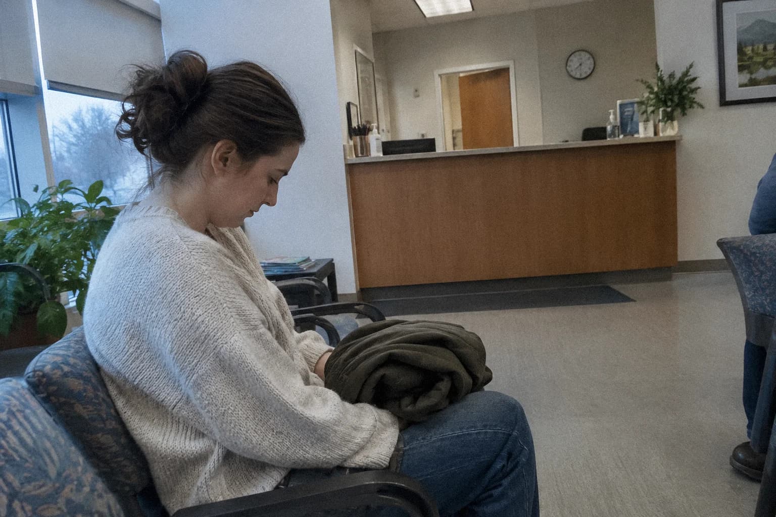 Une femme assise seule dans une salle d’attente, son manteau plié sur les genoux, le regard baissé vers le sol, le comptoir d’accueil vide derrière elle