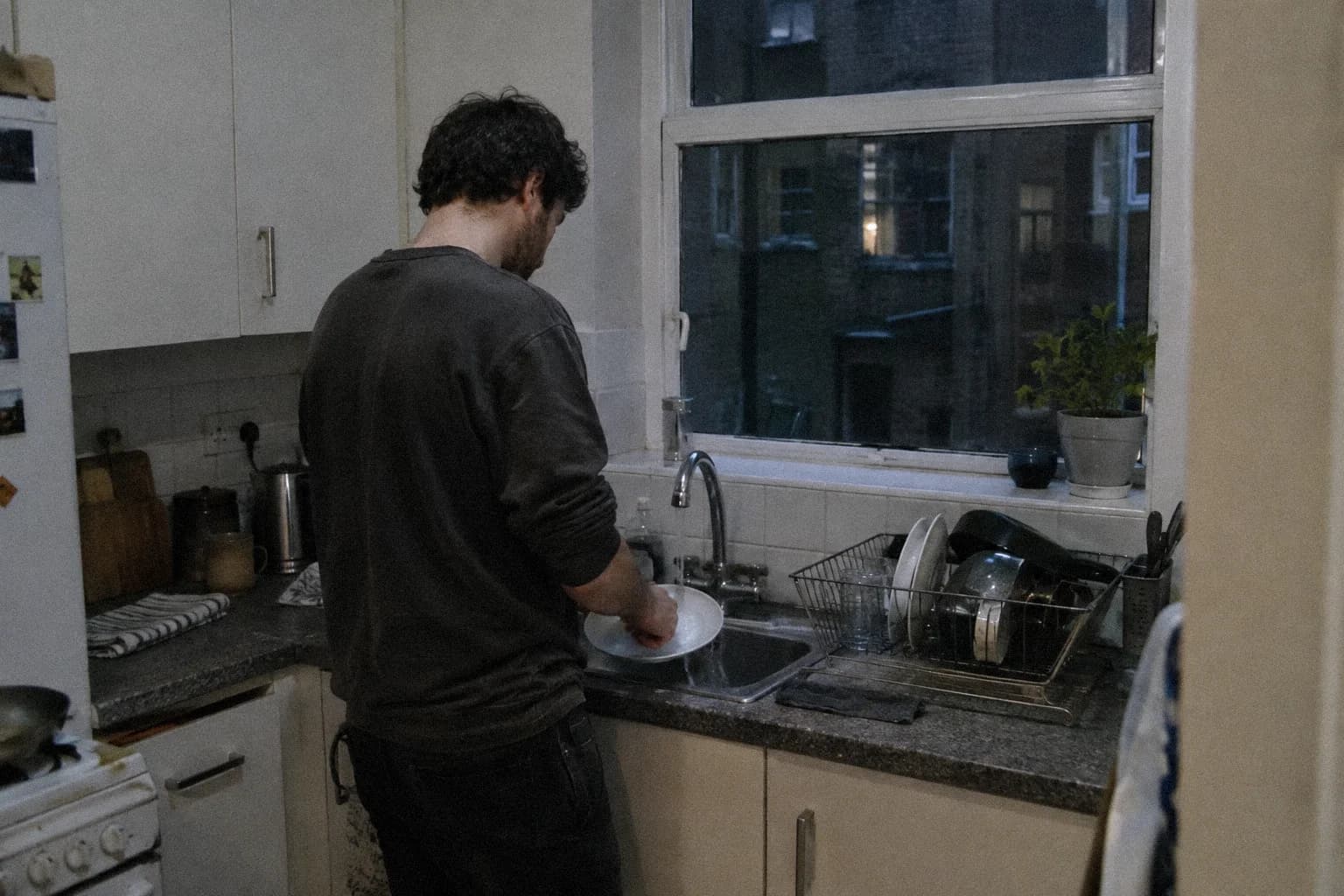 Un homme debout devant l’évier de sa cuisine tard le soir, lavant une assiette, la fenêtre donnant sur une cour sombre