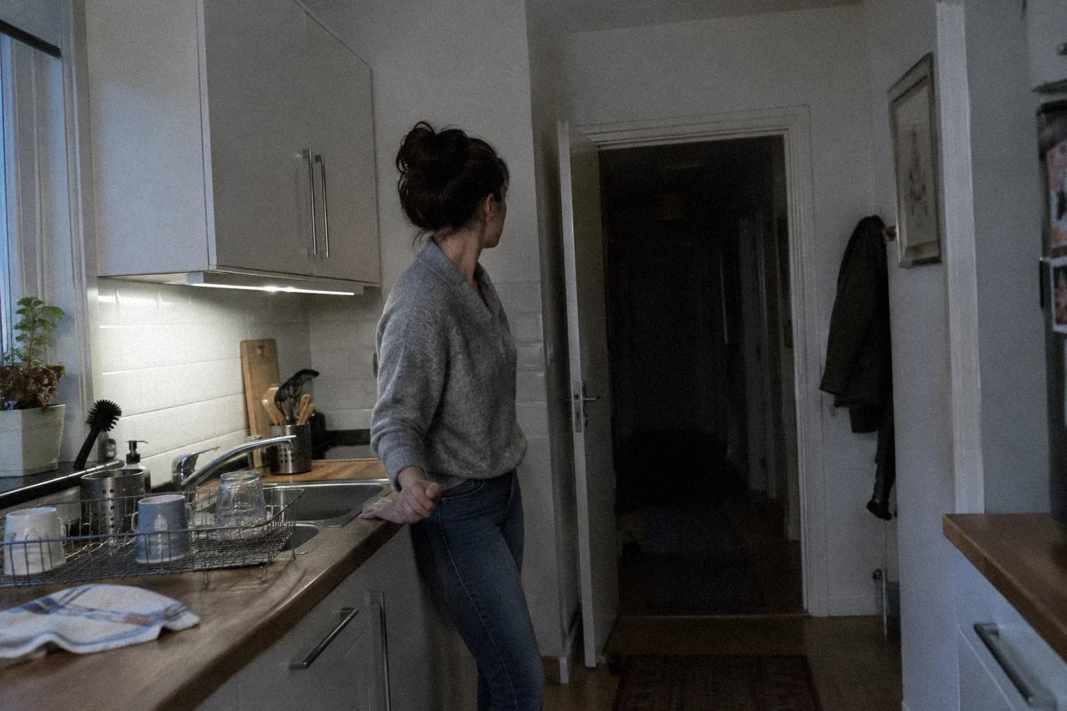 Une femme immobile devant l’évier de sa cuisine la nuit, la tête tournée vers le couloir sombre, les épaules remontées