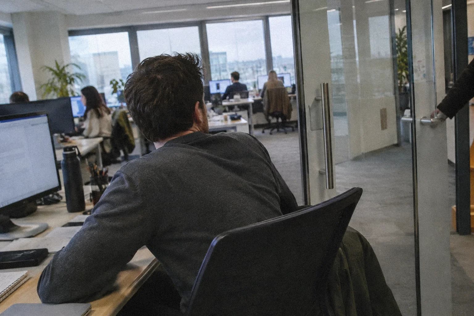 Un homme de dos à son bureau dans un open space, les épaules contractées, la tête à demi tournée vers une porte vitrée qui se referme derrière lui