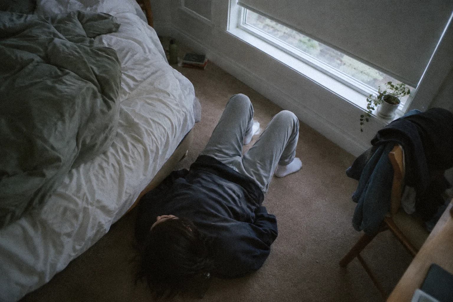 Vue de dessus d’une personne allongée sur la moquette d’une chambre à côté d’un lit défait, genoux relevés et pieds à plat, store à demi baissé
