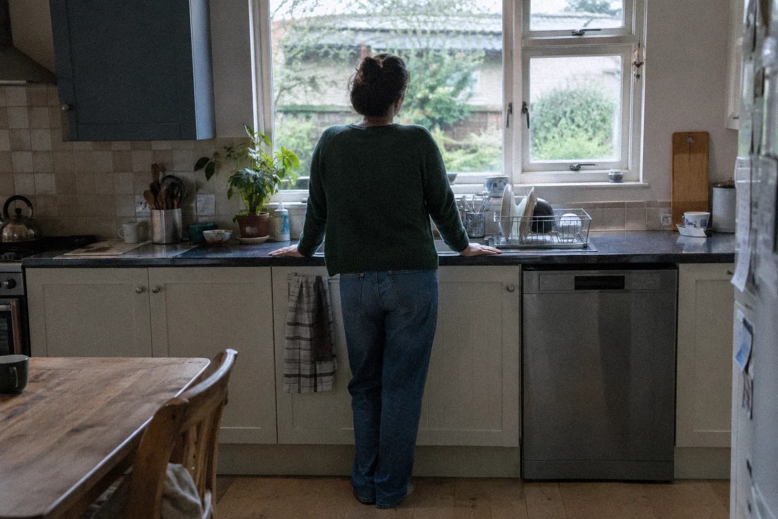 Une femme vue de dos, debout devant l’évier d’une cuisine, les mains posées sur le rebord du plan de travail, vaisselle sur l’égouttoir