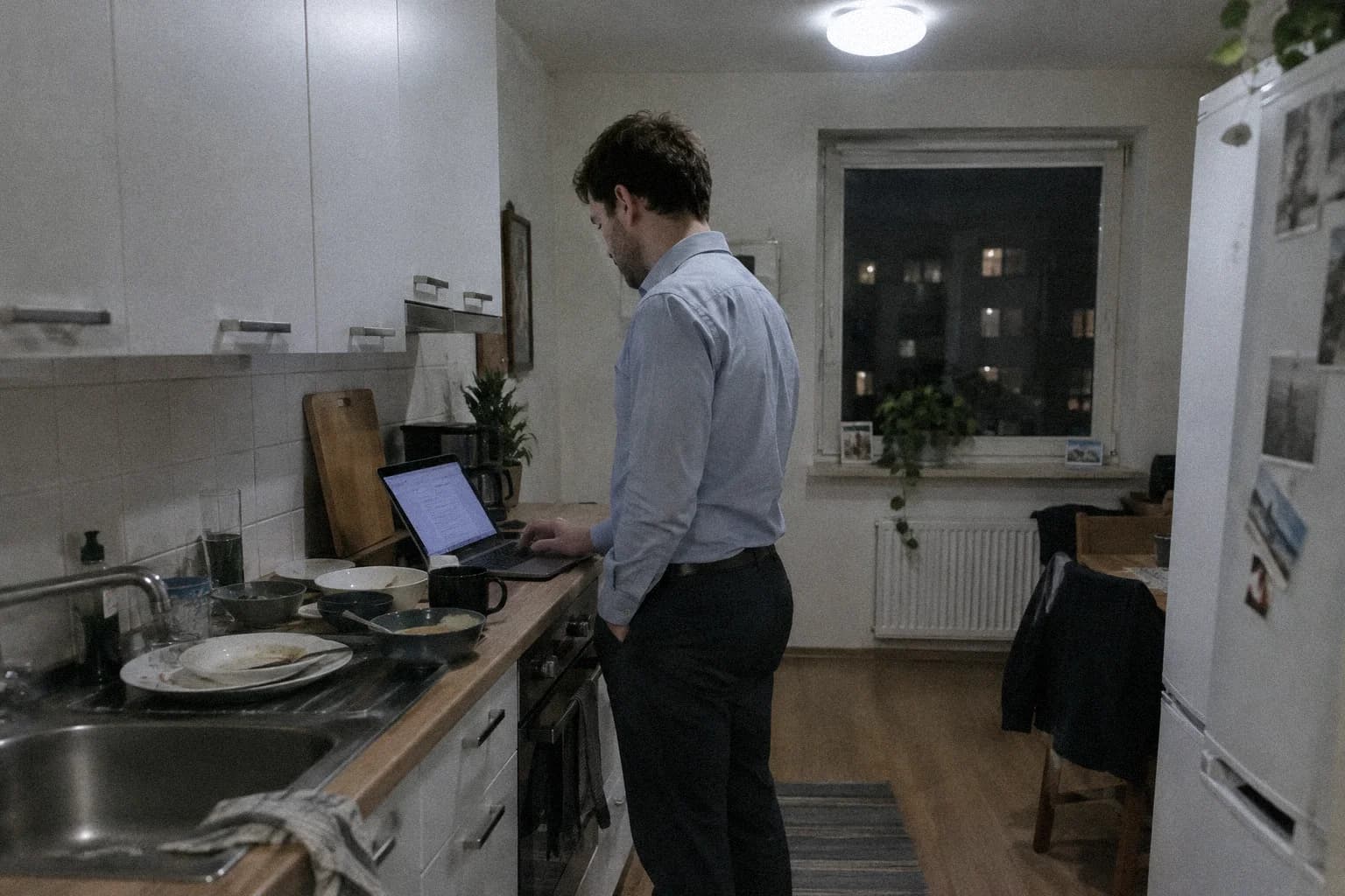 Arrêter de se mettre la pression : un homme debout dans sa cuisine tard le soir, encore en chemise de travail, relisant un document sur son ordinateur portable posé près de la vaisselle sale