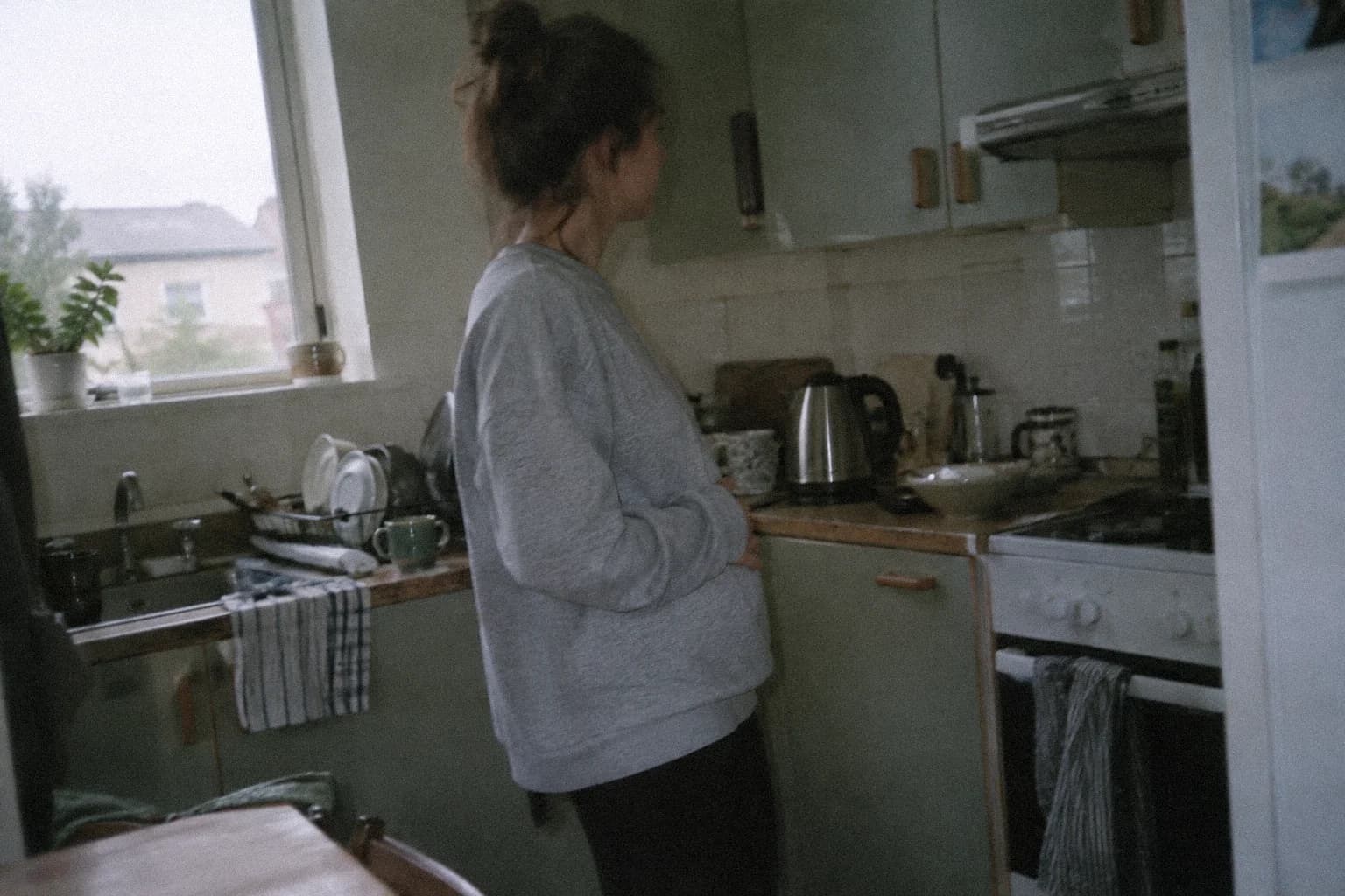 Une femme debout dans sa cuisine tôt le matin, de profil, les bras repliés devant le ventre