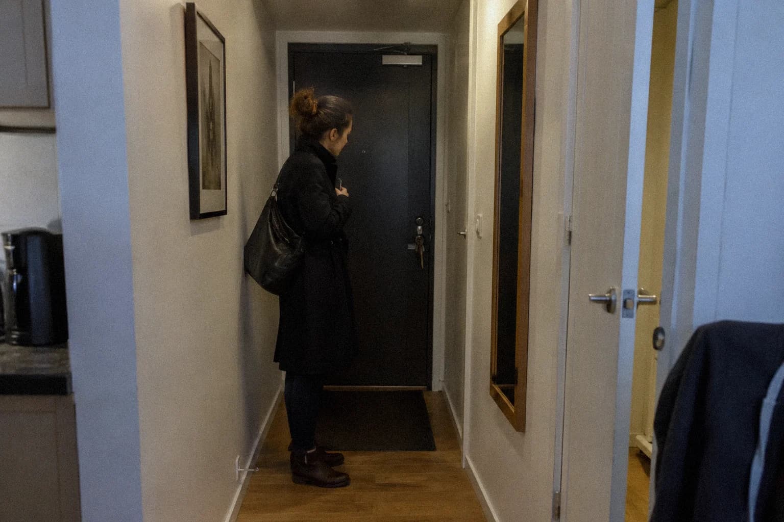 How to say no without feeling guilty: a woman standing in her apartment hallway in the evening, still in her coat, holding her phone face-down against her chest