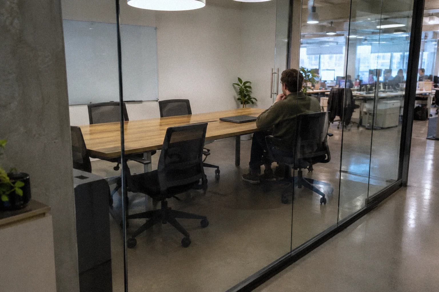 A man sitting alone in a glass-walled meeting room after the meeting, his laptop closed in front of him, the other chairs empty