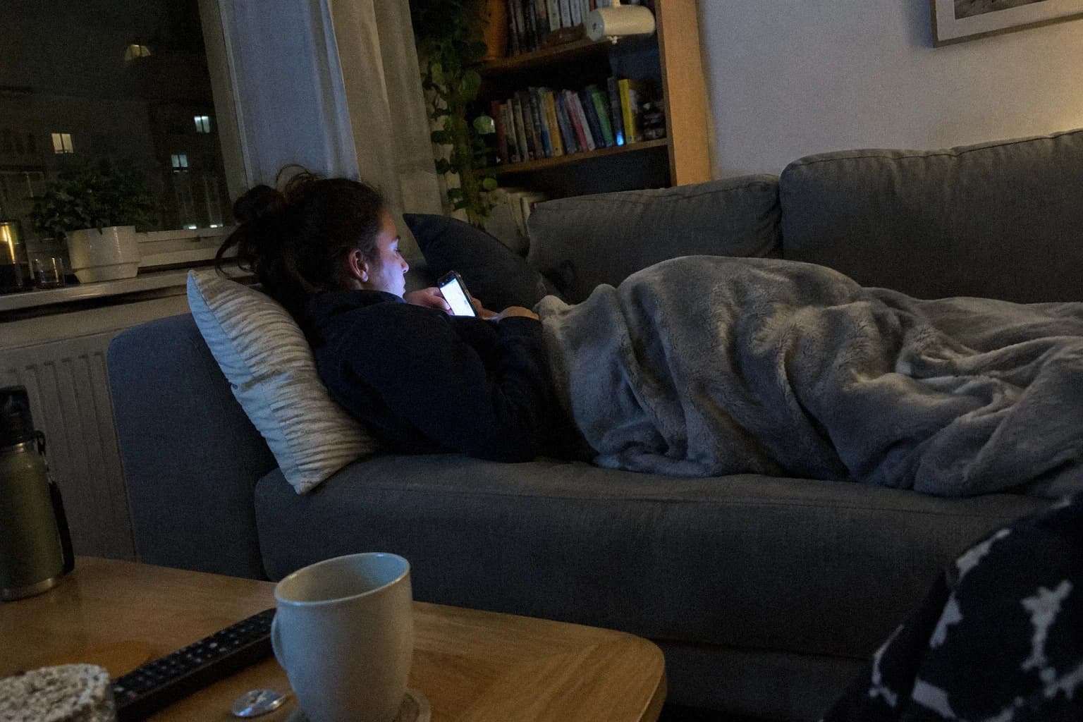 A woman lying on her sofa in a dark living room, phone in hand, the screen lighting her face, a blanket folded over her legs