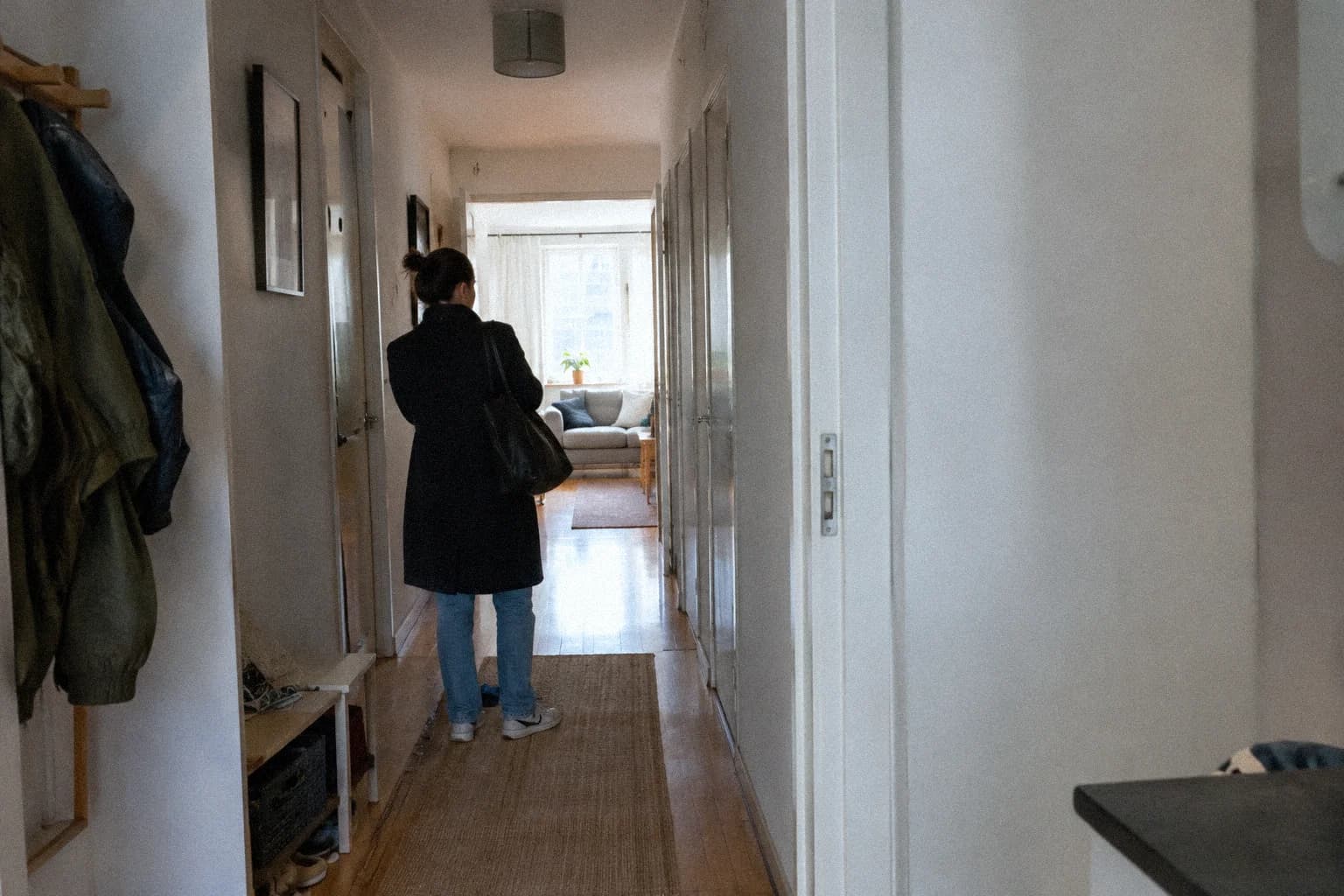How to switch off from work in the evening: a woman standing with her back to us in her apartment hallway, laptop bag still on her shoulder, the living room lit at the end of the corridor