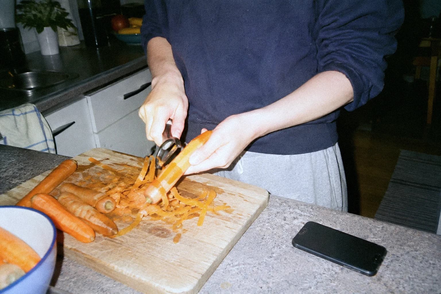 Someone peeling carrots in a kitchen in the evening, the cutting board covered in peelings, a phone lying face down on the counter