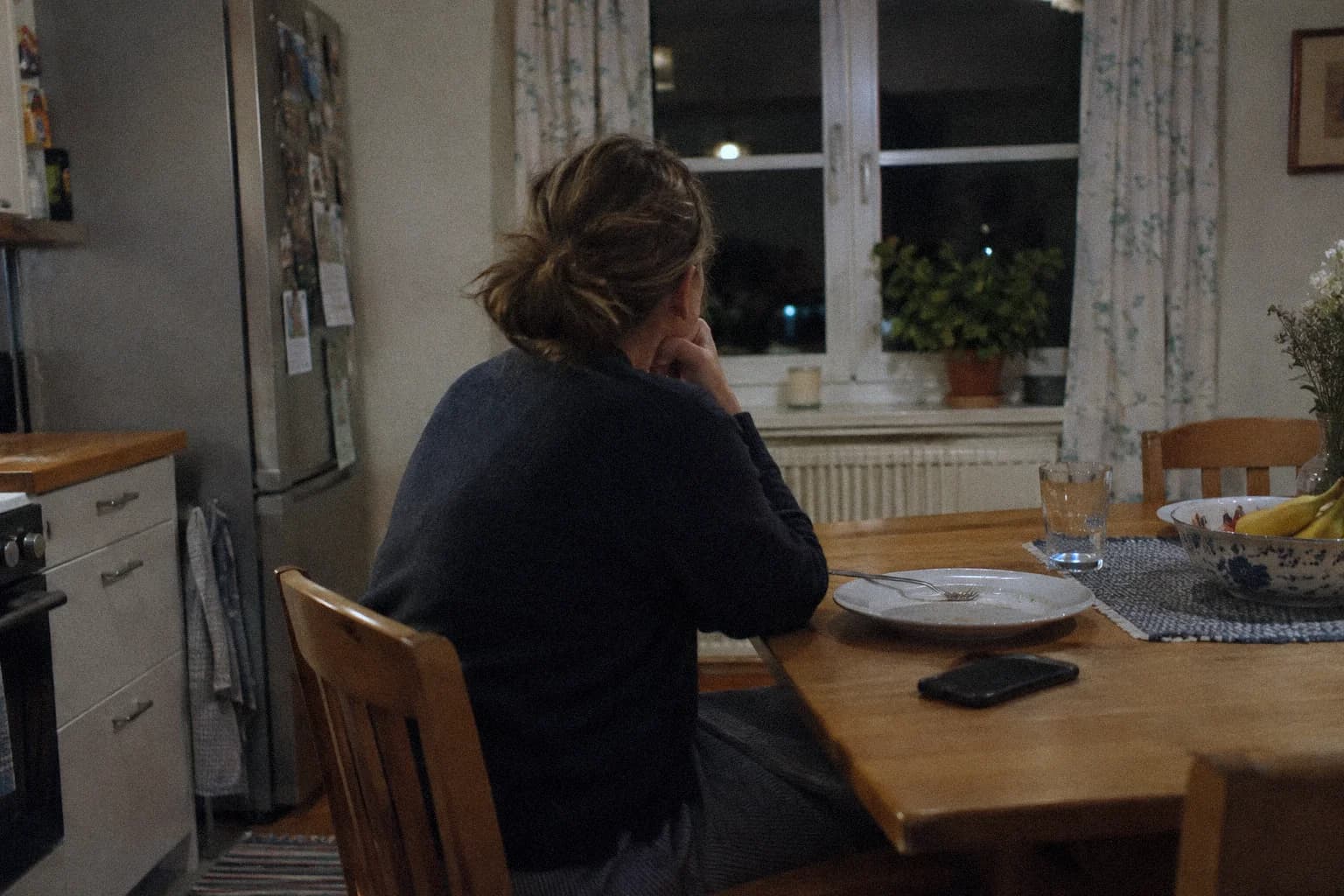 A woman sitting alone at a kitchen table in the evening, looking past the window, a finished plate and a face-down phone in front of her, after forgiving without the pain easing