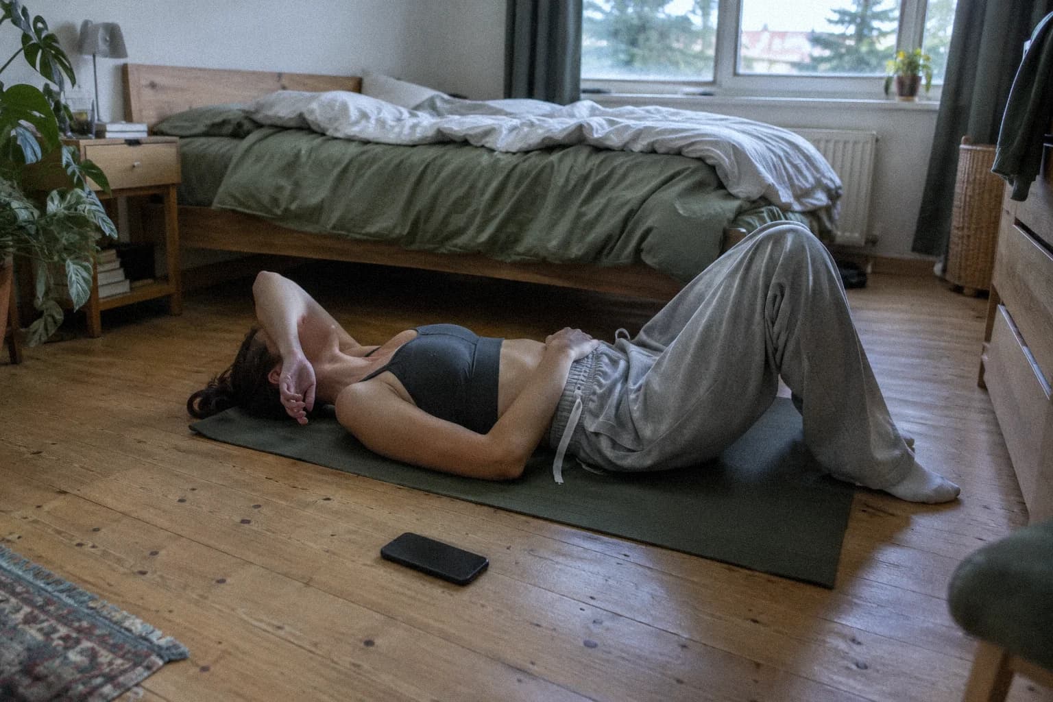 Une femme allongée sur le dos sur un tapis de sol dans une chambre en fin d’après-midi, un avant-bras tombé sur les yeux, un téléphone retourné sur le parquet à côté d’elle