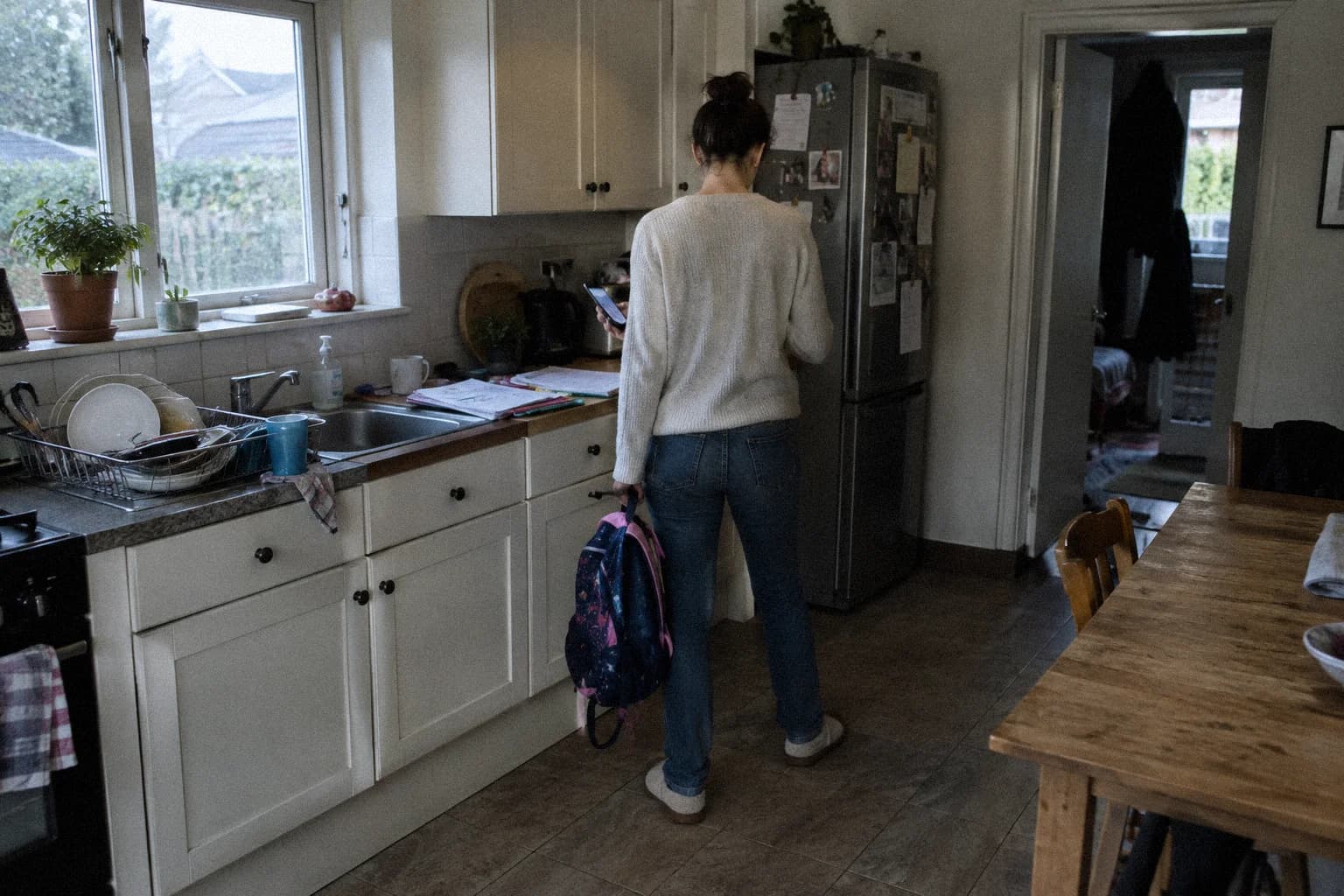 A woman standing with her back to the camera in a cluttered family kitchen in the evening, a phone in one hand and a child's backpack in the other, dishes still in the sink