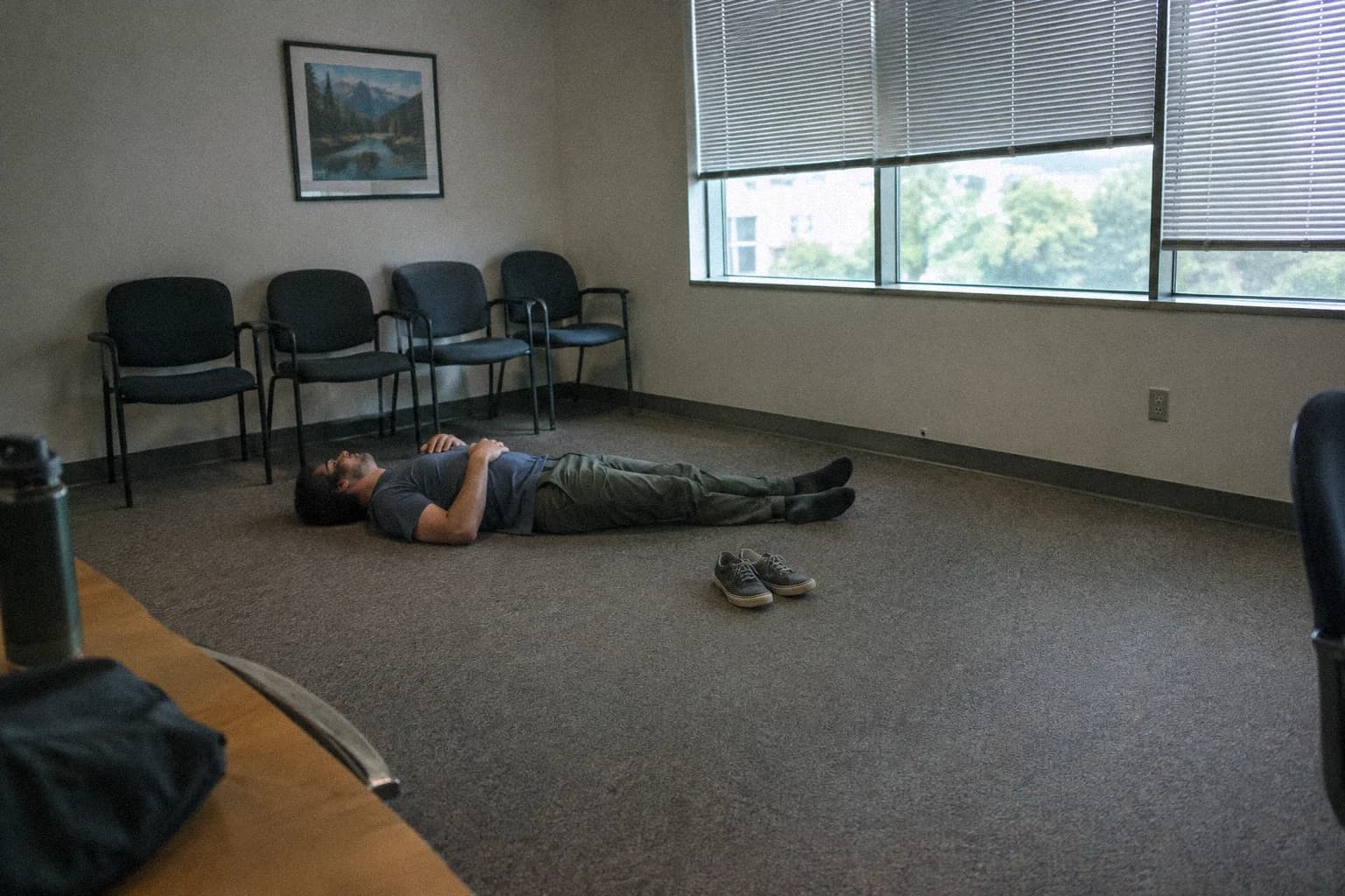 A man lying on his back on the carpet of a small empty meeting room at lunchtime, shoes off beside him, chairs pushed against the wall