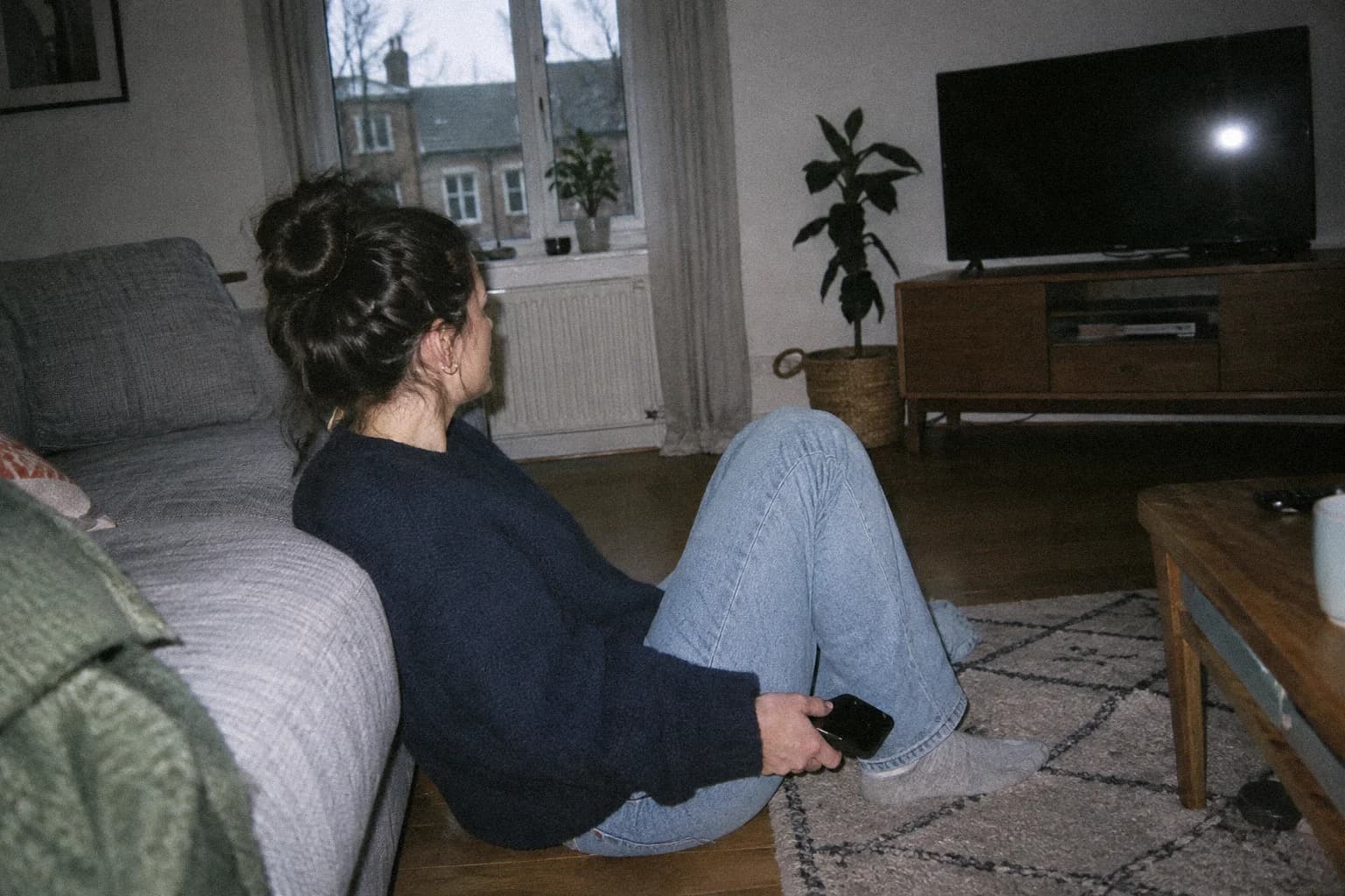 Femme assise par terre dos au canapé dans un petit salon, télévision éteinte, lumière grise d’après-midi à la fenêtre