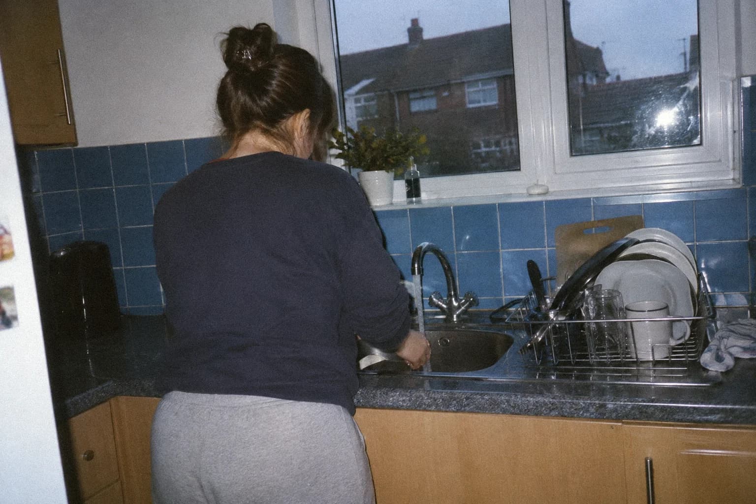 Femme debout devant un évier de cuisine, robinet ouvert, vue de dos, carrelage bleu et fenêtre au-dessus de l’évier