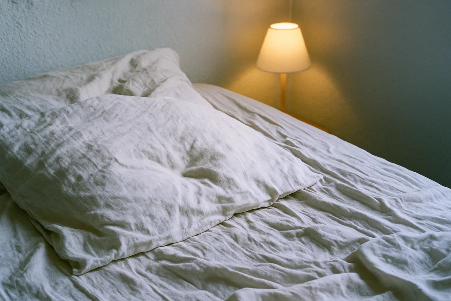 Un lit défait, oreiller de lin froissé et lampe de chevet allumée, quand le corps reste tendu au repos