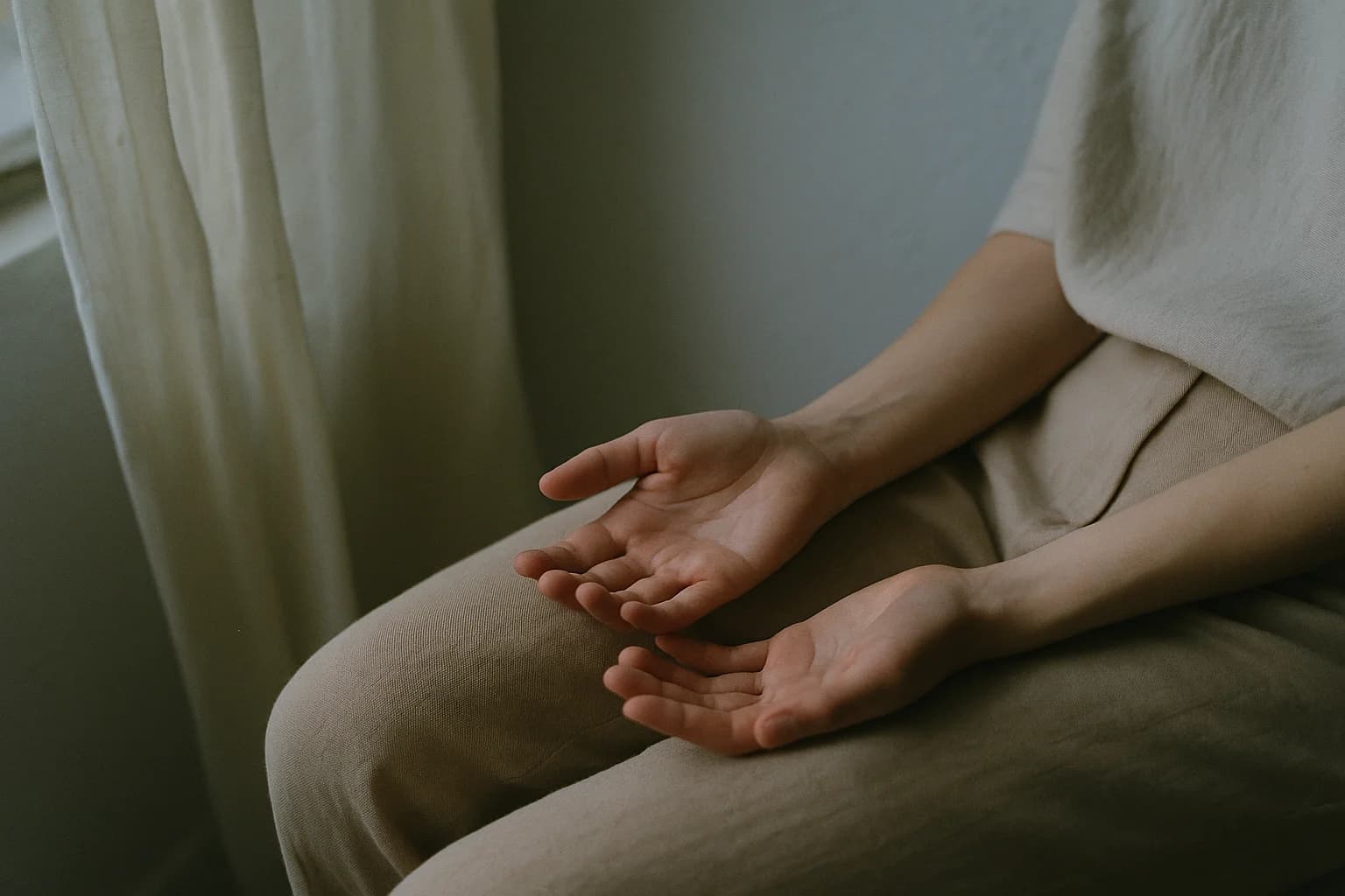 Deux mains posées paumes ouvertes sur les cuisses d'une personne assise, doigts détendus, lumière de fenêtre venant de la gauche