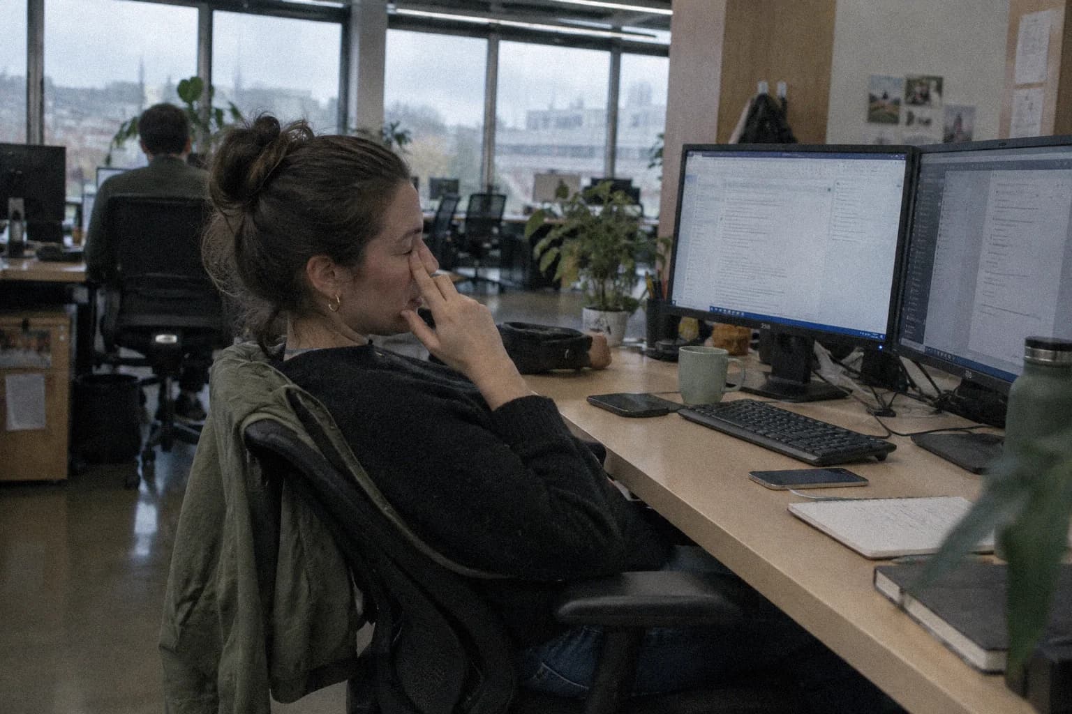 Eye twitching from stress: a woman at her open-plan office desk late in the day, one eye shut with two fingers pressed under it, two monitors still on in front of her