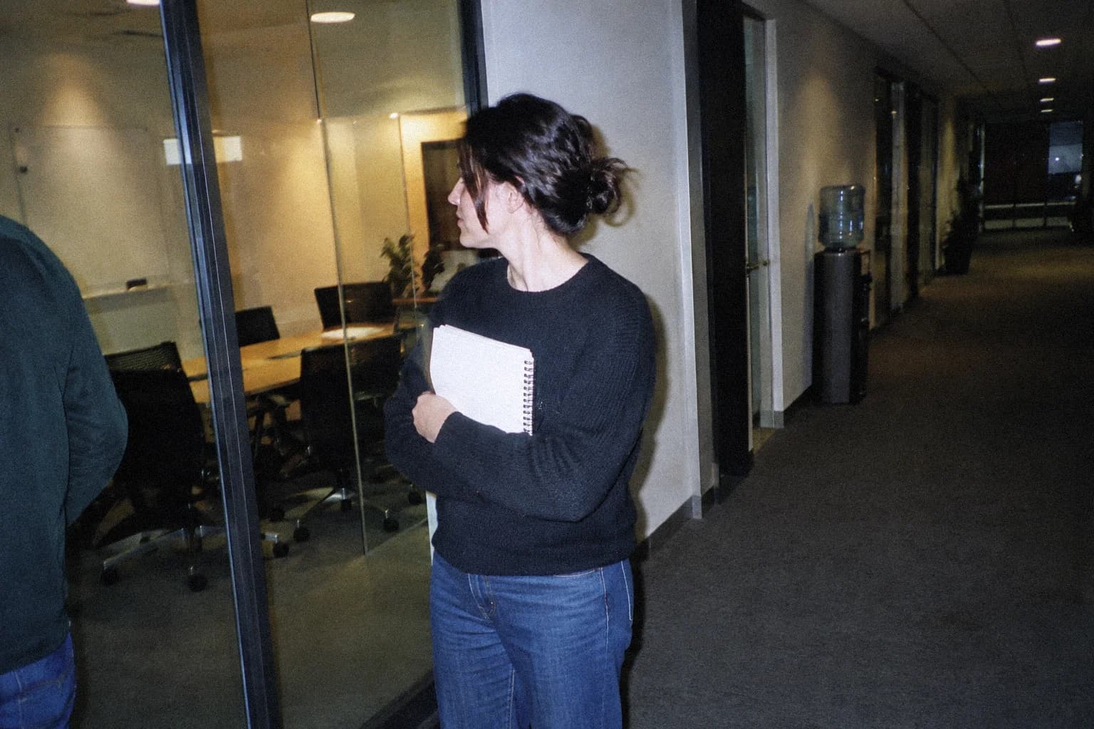 Une femme debout dans un couloir de bureau après une réunion, son carnet serré contre elle, la cloison vitrée de la salle de réunion derrière elle et une fontaine à eau plus loin dans le couloir