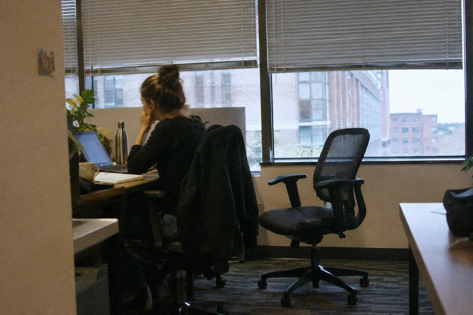 A woman sitting at her desk late in the day, an empty chair beside her, office blinds half lowered