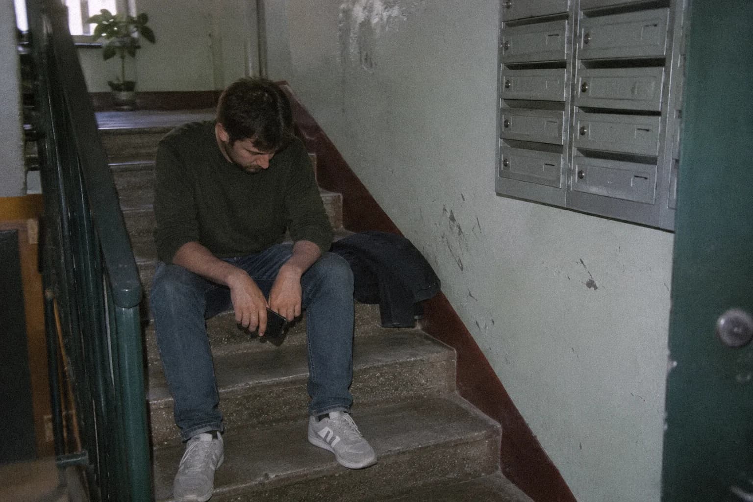 How to rebuild confidence after failure: a man sitting on the concrete steps of an apartment stairwell, his jacket folded beside him, phone loose in one hand