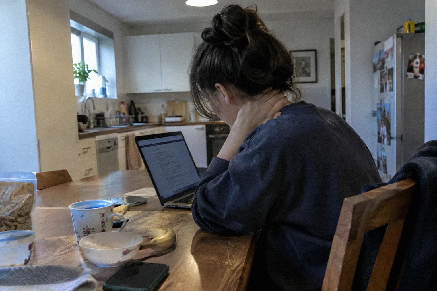 A woman at a kitchen table in the morning in front of an open laptop, shoulders rounded forward, one hand on the back of her neck