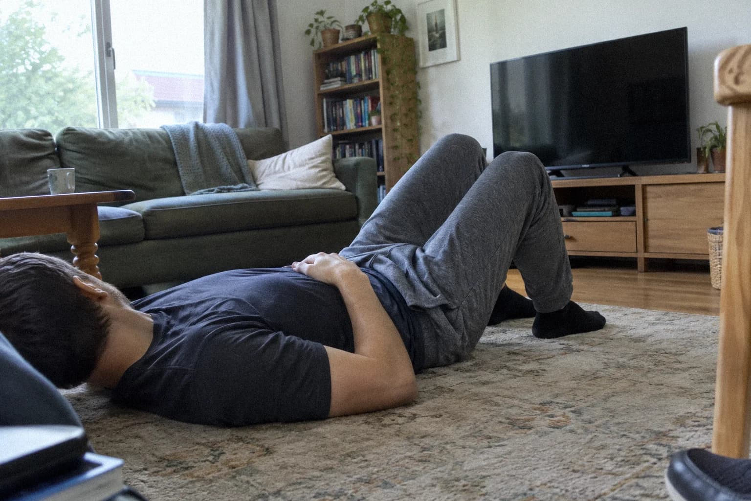 A man lying on his back on a living room rug, knees bent and feet flat, one hand resting on his stomach, a sofa and a switched-off television behind him