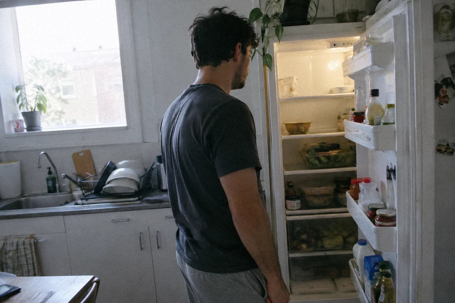 Retrouver le plaisir de vivre : un homme debout devant son réfrigérateur ouvert le soir, dans une cuisine éclairée par la seule lampe de l’appareil, regardant les étagères sans rien prendre