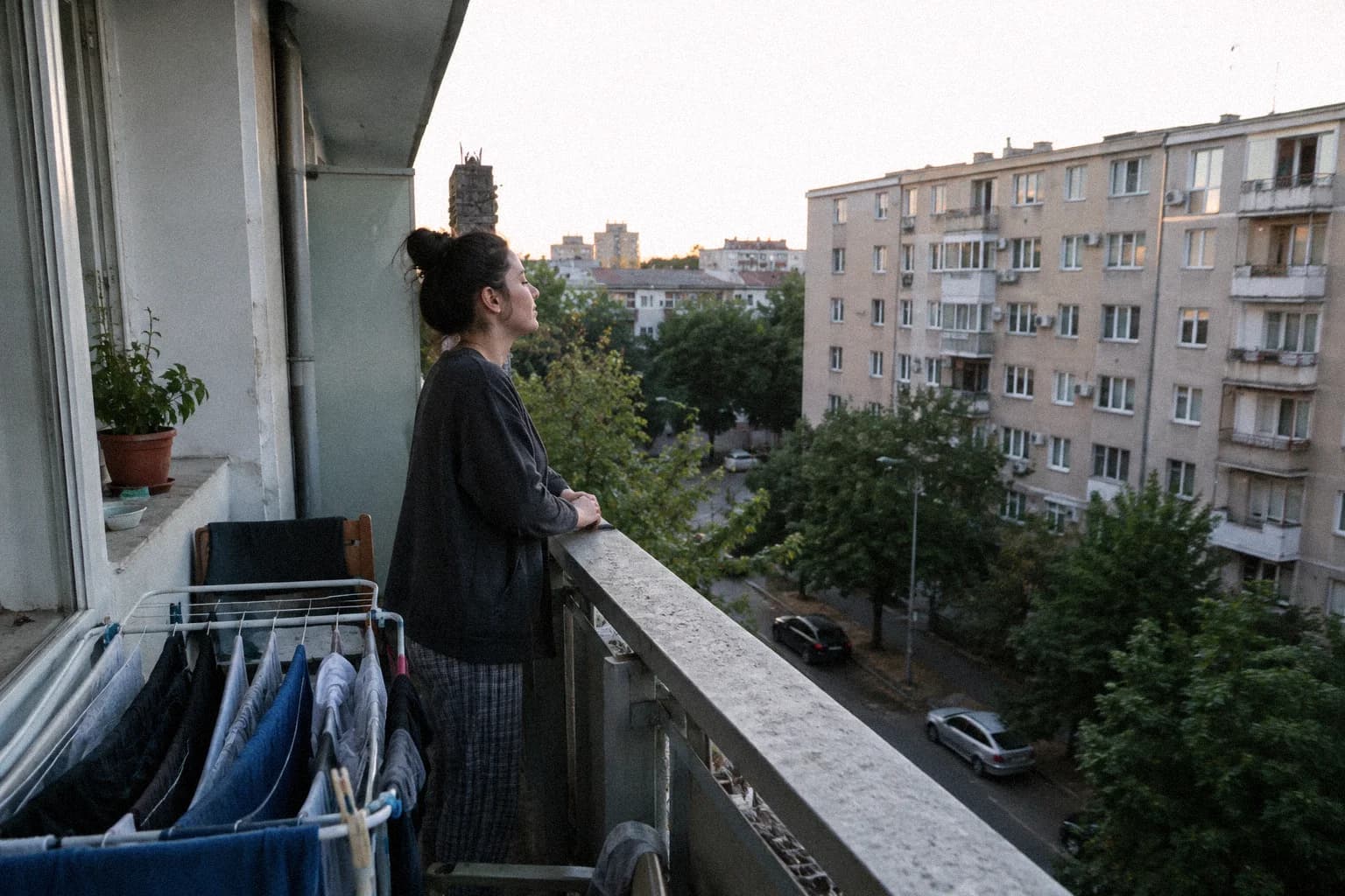 Une femme debout sur le balcon d’un immeuble à la tombée du jour, accoudée à la rambarde, écoutant la rue en contrebas, un étendoir à linge derrière elle