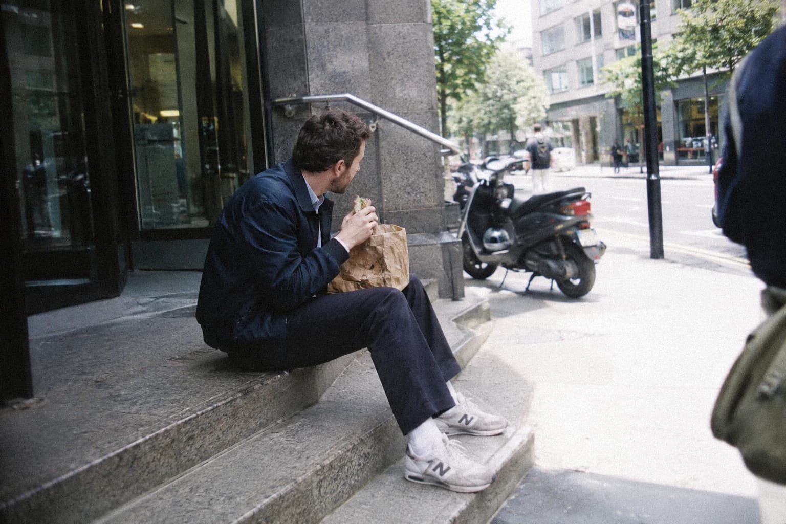 Un homme assis sur les marches de l’entrée d’un immeuble de bureaux, mangeant un sandwich pendant sa pause déjeuner, un scooter garé le long du trottoir