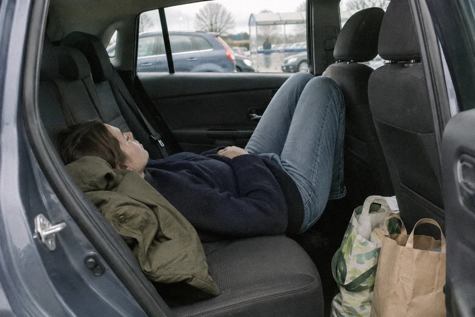 A woman lying across the back seat of a car parked in a supermarket car park, knees bent, a folded coat under her head, shopping bags on the floor