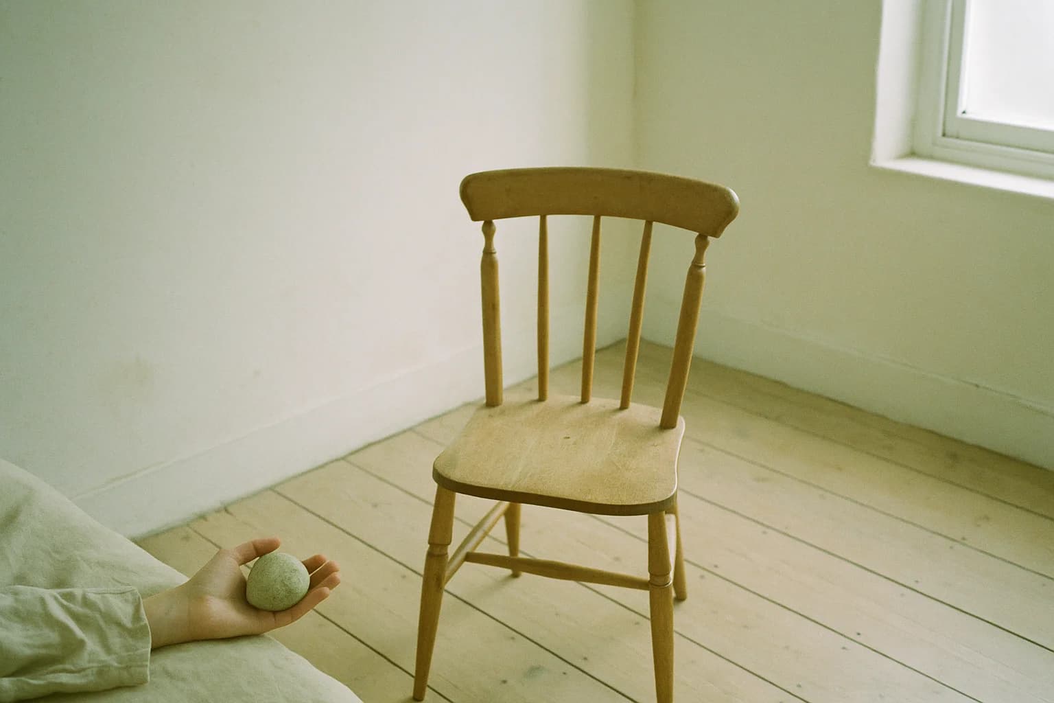 Chaise en bois vide dans une pièce claire aux lames de parquet larges, une main au sol tenant un galet